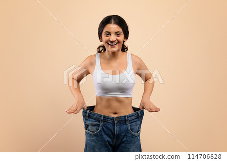 Weight Loss Result. Happy Young Indian Woman Posing In Oversized Jeans, Slender Female Wearing Big Clothes Standing On Beige Studio Background, Comparing Sizes After Successful Diet, Copy Space Weight Loss Result. Happy Young Indian Woman Posing In Oversized Jeans, Slender Female Wearing Big Clothes Standing On Beige Studio Background, Comparing Sizes After Successful Diet, Copy Space 114706828