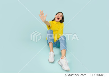 Joyful young brunette woman wearing stylish casual outfit sitting on floor and laughing, waving at camera, isolated on blue studio background, copy space 114706830
