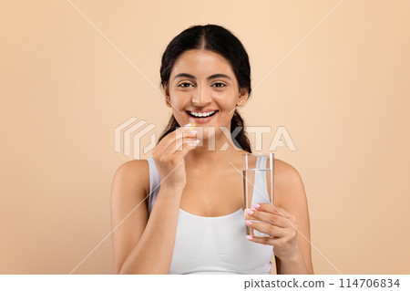 Beauty Supplement. Attractive Indian Woman Holding Vitamin Capsule And Water Glass, Smiling Eastern Female Taking Pill For Beautiful Skin, Hair And Nails, Standing Isolated On Beige Background Beauty Supplement. Attractive Indian Woman Holding Vitamin Capsule And Water Glass, Smiling Eastern Female Taking Pill For Beautiful Skin, Hair And Nails, Standing Isolated On Beige Background 114706834