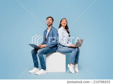 Young European spouses seated back to back, deeply engrossed in their laptops and glancing upwards at free space on soothing blue studio background, banner Young European spouses seated back to back, deeply engrossed in their laptops and glancing upwards at free space on soothing blue studio background, banner 114706835