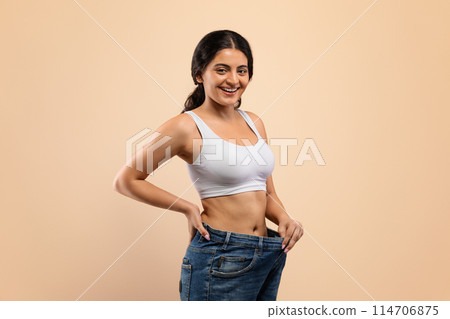 Happy Young Indian Woman Wearing Oversized Jeans Demonstrating Weight Loss Result, Smiling Eastern Female Comparing Sizes After Successful Diet, Posing Over Beige Background In Studio, Copy Space 114706875