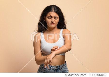 Skin Disorders. Young indian woman suffering eczema, scratching irritated skin on arm, upset eastern female having itching rash on body, standing isolated on beige studio background, copy space Skin Disorders. Young indian woman suffering eczema, scratching irritated skin on arm, upset eastern female having itching rash on body, standing isolated on beige studio background, copy space 114706889