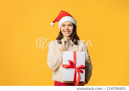 Millennial lady with joyful expression holds Christmas present, ready to celebrate and give gift, standing against yellow studio setting, touching chin with smile. New year season, festive offer 114706891