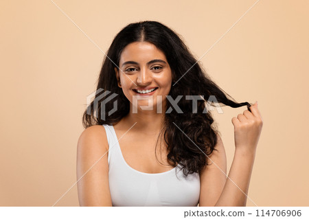Happy young indian female showing her beautiful long hair after salon treatment and smiling at camera, attractive eastern female touching curl, standing isolated over beige studio background 114706906