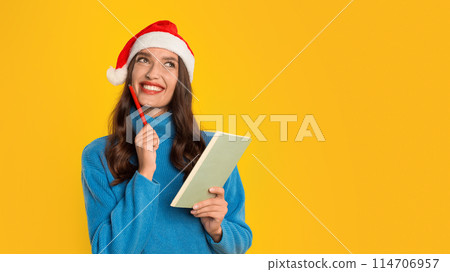 European woman smiles dreaming and writing down Christmas festive dreams and sales wishes in her notebook, standing on yellow studio background. Panorama, free space for text 114706957
