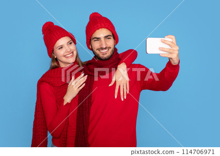 Winter Selfie. Happy Young Couple In Knitted Hats Making Photos On Smartphone, Cheerful Man And Woman Embracing While Posing At Mobile Phone Camera, Standing Isolated On Blue Background 114706971