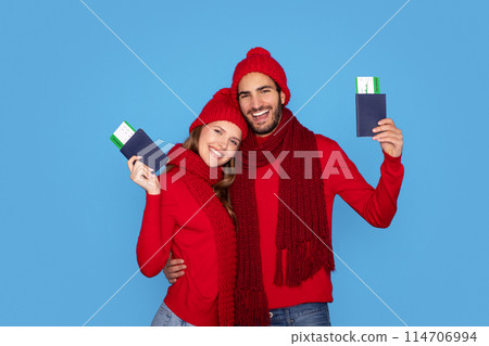 Travelling Concept. Young Smiling Couple Wearing Knitted Hats Holding Passports With Boarding Pass Tickets, Happy Man And Woman Ready For Winter Vacation, Posing Isolated On Blue Studio Background 114706994