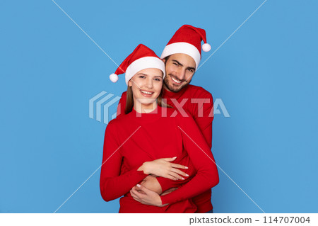 Happy young couple wearing Santa hats embracing and smiling at camera, cheerful european man and woman posing together on festive blue studio background, enjoying Christmas holidays celebration Happy young couple wearing Santa hats embracing and smiling at camera, cheerful european man and woman posing together on festive blue studio background, enjoying Christmas holidays celebration 114707004
