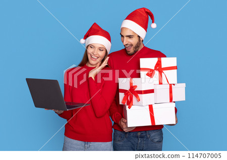 Online Shopping. Excited Young Couple Wearing Santa Hats Holding Gifts And Using Laptop, Cheerful Man And Woman Buying Presents In Internet, Standing Isolated On Blue Studio Background, Free Space Online Shopping. Excited Young Couple Wearing Santa Hats Holding Gifts And Using Laptop, Cheerful Man And Woman Buying Presents In Internet, Standing Isolated On Blue Studio Background, Free Space 114707005