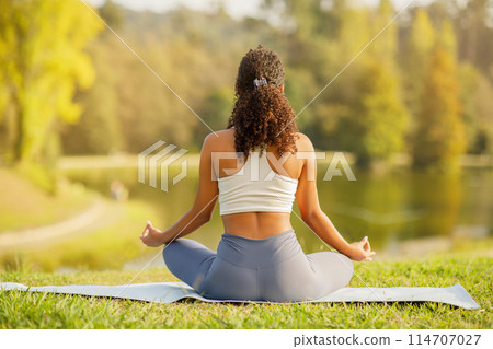 Young sporty woman meditating in lotus position on mat by the lake, rear view, epitome of calmness and wellness, ideal for yoga and fitness ad, outdoors in park on summer morning Young sporty woman meditating in lotus position on mat by the lake, rear view, epitome of calmness and wellness, ideal for yoga and fitness ad, outdoors in park on summer morning 114707027