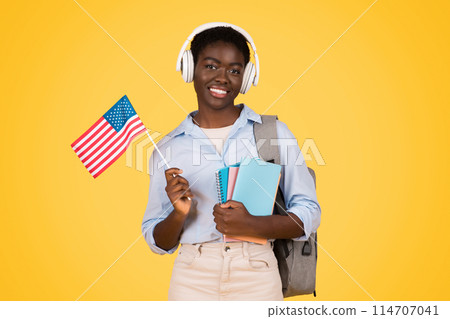Happy young african american woman student in wireless headphones, with books, flag of USA, listen music, isolated on yellow studio background. Study and education exchange, learn English Happy young african american woman student in wireless headphones, with books, flag of USA, listen music, isolated on yellow studio background. Study and education exchange, learn English 114707041