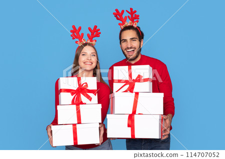 Cheerful Man And Woman Wearing Reindeer Party Antlers Horns Holding Stack Of Christmas Gift Boxes, Happy Couple Carrying Presents And Smiling At Camera, Posing Isolated On Blue Background, Copy Space 114707052
