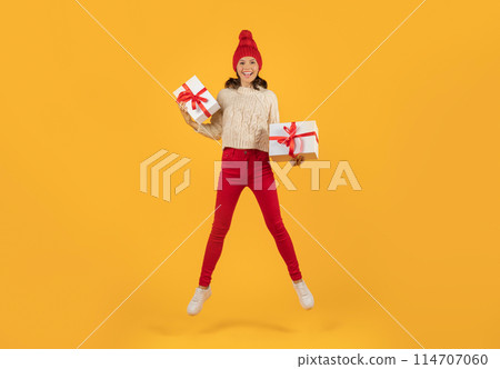 Cheerful young woman in red knitwear, joyfully jumping with two Christmas gifts boxes, bright and festive, studio shot against yellow background, perfect for Xmas holiday sale banners Cheerful young woman in red knitwear, joyfully jumping with two Christmas gifts boxes, bright and festive, studio shot against yellow background, perfect for Xmas holiday sale banners 114707060