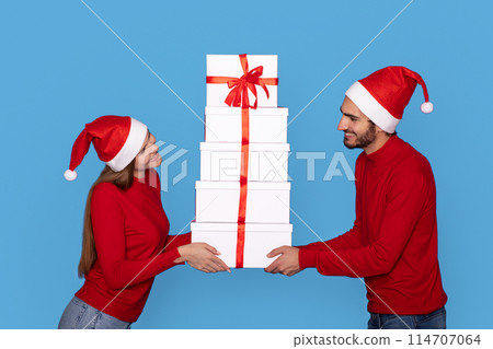 Young Happy Couple Handling Stack Of Gift Boxes With Red Ribbons, Cheerful Man And Woman Wearing Santa Hats Enjoying Christmas Present Exchanges And Holidays Celebrations, Standing On Blue Background 114707064