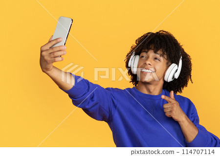Happy young black guy wearing wireless headphones taking selfie with smartphone, cheerful handsome african american man showing gotcha gesture at mobile phone camera, standing on yellow background 114707114