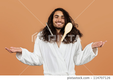 Carefree Guy In Bathrobe Posing With Hairbrush And Comb Stuck In His Thick Curly Hair, Shrugging And Smiling While Having Problem With Detangling Of Long Brunette Hair, On Beige Background 114707126