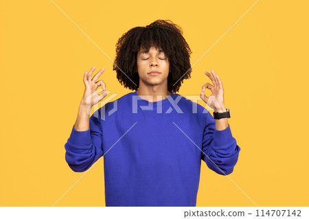 Serene black man in blue sweatshirt meditating with closed eyes, calm african american guy making ok sign with fingers while standing isolated on yellow background, embodying mindfulness, copy space 114707142