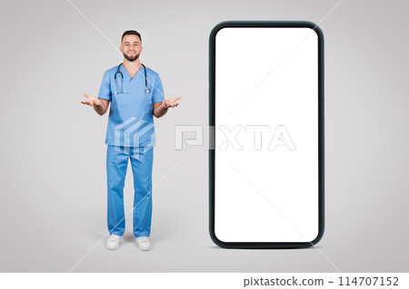 Cheerful male nurse in blue scrubs standing next to an oversized smartphone, gesturing towards the blank screen, representing telehealth services, mockup 114707152