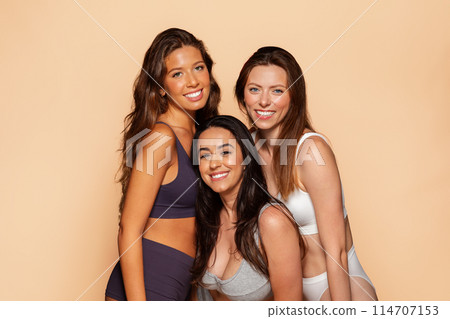 Three happy young international women in casual underwear together, smiling, embracing from behind, isolated on beige background. Friendship and togetherness, spa, beauty care 114707153