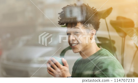 Shot from outside of happy stylish young black guy drinking coffee at cafe, holding green mug and looking at copy space, panorama. Leisure, millennials lifestyle concept Shot from outside of happy stylish young black guy drinking coffee at cafe, holding green mug and looking at copy space, panorama. Leisure, millennials lifestyle concept 114707233