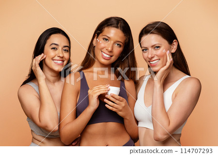 Three multiethnic young women in underwear show jar, put cream on face, enjoy beauty care, hydration at home on beige studio background. Fun lifestyle, cosmetics and spa, close up Three multiethnic young women in underwear show jar, put cream on face, enjoy beauty care, hydration at home on beige studio background. Fun lifestyle, cosmetics and spa, close up 114707293