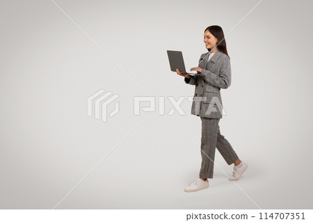 Active young professional woman walking and using laptop, dressed in houndstooth suit with casual sneakers, against minimalistic grey backdrop, free space 114707351