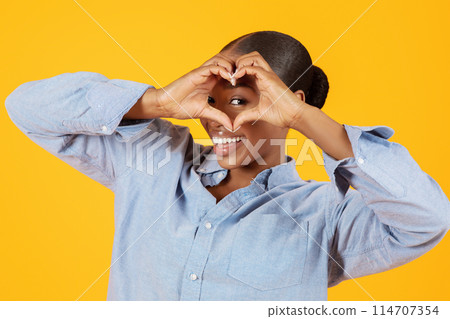 Funny African American Young Lady Looking At Camera Through Heart Shaped Fingers Gesture, Over Yellow Studio Backdrop, Flirting And Advertising Dating Service. I Like It Concept Funny African American Young Lady Looking At Camera Through Heart Shaped Fingers Gesture, Over Yellow Studio Backdrop, Flirting And Advertising Dating Service. I Like It Concept 114707354