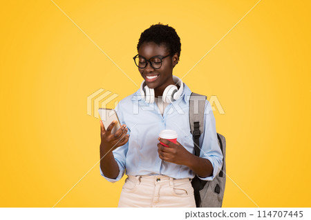 Glad black woman student in glasses, multitasking with coffee in hand and phone, isolated on yellow background. Study, busy lifestyle, chat in social networks, break and education Glad black woman student in glasses, multitasking with coffee in hand and phone, isolated on yellow background. Study, busy lifestyle, chat in social networks, break and education 114707445