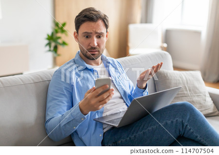 A man sits on a couch with a laptop on his thighs, holding a smartphone in one hand while displaying a puzzled expression, appears to be encountering unexpected news or a perplexing situation 114707504