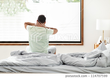 Black man sits on a neatly made bed with his back to the camera, hands clasped behind his head, stretching while looking out of a large bedroom window. The morning light fills the room Black man sits on a neatly made bed with his back to the camera, hands clasped behind his head, stretching while looking out of a large bedroom window. The morning light fills the room 114707599