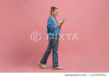 A young girl wearing a blue shirt is focused on her cell phone screen in this candid moment captured in a public setting. She appears engrossed in her phone, possibly messaging or scrolling A young girl wearing a blue shirt is focused on her cell phone screen in this candid moment captured in a public setting. She appears engrossed in her phone, possibly messaging or scrolling 114707612