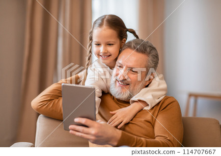 An older man and a young girl are sitting side by side on a couch, both engrossed in the content on a tablet. They appear focused and engaged An older man and a young girl are sitting side by side on a couch, both engrossed in the content on a tablet. They appear focused and engaged 114707667
