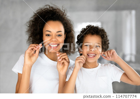 A cheerful African American mother and her young son are standing together in a well-lit, contemporary bathroom, wearing white t-shirts while they both floss their teeth 114707774