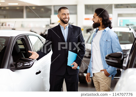 A smartly dressed car salesman, with a friendly demeanor, stands next to a shiny new car in a well-lit vehicle showroom, engaging with a potential customer Indian guy who is holding a brochure. A smartly dressed car salesman, with a friendly demeanor, stands next to a shiny new car in a well-lit vehicle showroom, engaging with a potential customer Indian guy who is holding a brochure. 114707776