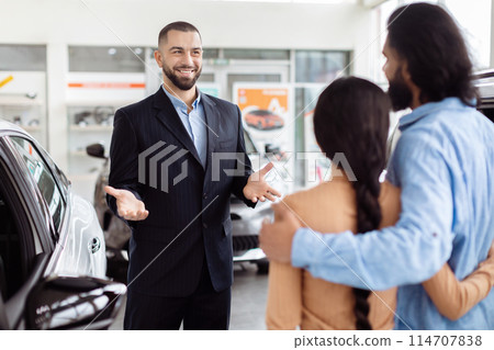 A cheerful car salesman is having a conversation with Indian couple inside a car dealership showroom, he seems to be explaining the features or benefits of a vehicle A cheerful car salesman is having a conversation with Indian couple inside a car dealership showroom, he seems to be explaining the features or benefits of a vehicle 114707838