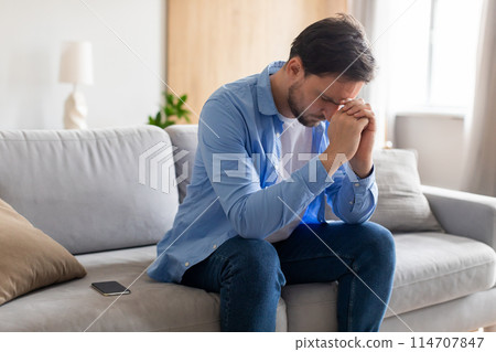A man is seated on a couch with his hands covering his face, appearing deep in thought or distressed. He looks contemplative and lost in his own thoughts. A man is seated on a couch with his hands covering his face, appearing deep in thought or distressed. He looks contemplative and lost in his own thoughts. 114707847