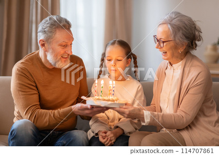 A young girl sitting between her smiling grandparents is blowing out candles on a birthday cake that her grandfather is holding. They appear to be seated comfortably in a living room A young girl sitting between her smiling grandparents is blowing out candles on a birthday cake that her grandfather is holding. They appear to be seated comfortably in a living room 114707861