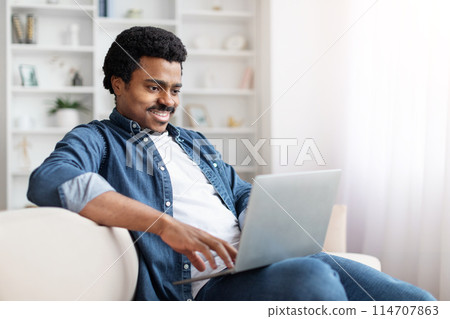 African American man is sitting on a couch, focused on using a laptop computer. Guy hands are typing on the keyboard, and the screen displays digital content. The room appears cozy and comfortable. African American man is sitting on a couch, focused on using a laptop computer. Guy hands are typing on the keyboard, and the screen displays digital content. The room appears cozy and comfortable. 114707863