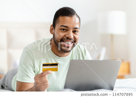 African American man is shown holding a credit card in one hand and a laptop in the other. He appears to be making a transaction or shopping online. 114707895