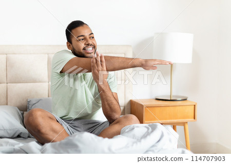 A young African American man is sitting on a bed, wearing casual sleepwear and stretching with a content smile 114707903