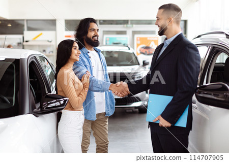 A young Indian couple is finalizing a deal with a car salesman inside a vehicle showroom. They seem pleased and are shaking hands with the agent, who is dressed in a business suit 114707905