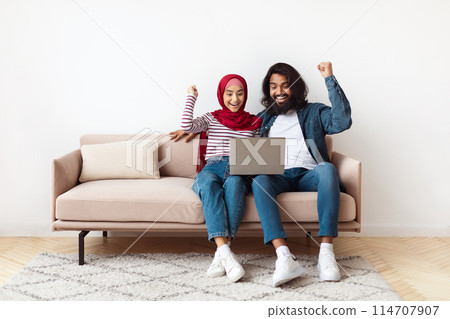 Eastern man and woman are seated on a couch, focused on a laptop screen in front of them. Both individuals appear engaged, possibly working or browsing the internet 114707907