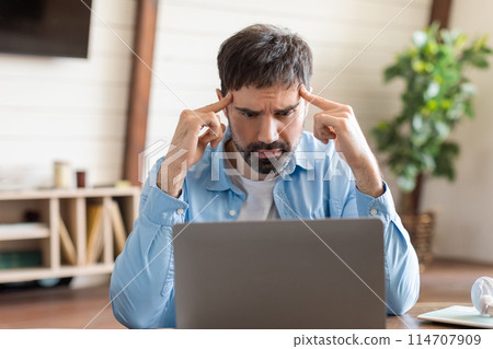 A man seated at a wooden desk, focusing intently on the contents of his laptop screen. His brows are furrowed, pressing his temples as if deep in thought or trying to solve a complex problem. 114707909