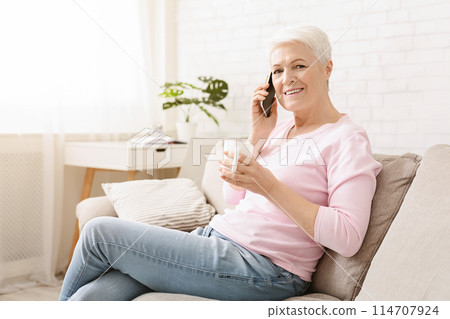 Senior woman is seated on a couch, engaged in conversation on her cell phone. She appears focused as she communicates with the person on the other end. 114707924