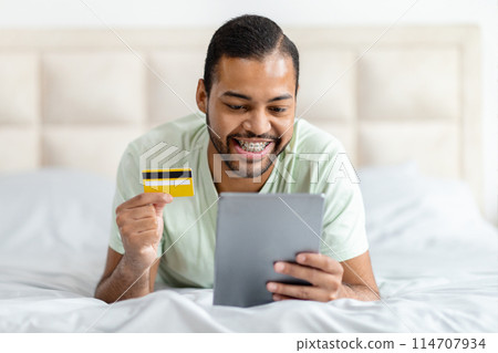 African American man is reclining on a bed while holding a credit card in one hand and a tablet in the other, possibly engaging in online shopping or managing finances. African American man is reclining on a bed while holding a credit card in one hand and a tablet in the other, possibly engaging in online shopping or managing finances. 114707934