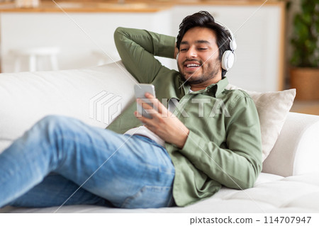 Arab man is seated on a couch, attentively listening to music playing from his phone, using headphones. He appears relaxed as he enjoys the music through his headphones. Arab man is seated on a couch, attentively listening to music playing from his phone, using headphones. He appears relaxed as he enjoys the music through his headphones. 114707947