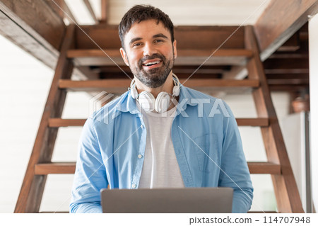 A man sitting with headphones on, focused on his laptop screen, typing and interacting with the digital device. He appears engaged in work or leisure activities. A man sitting with headphones on, focused on his laptop screen, typing and interacting with the digital device. He appears engaged in work or leisure activities. 114707948