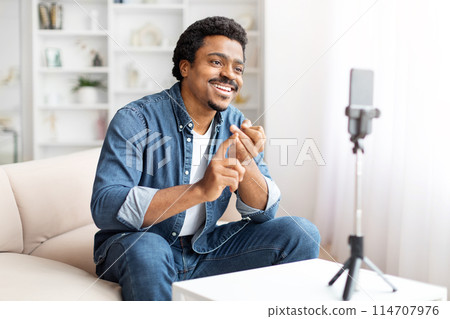 A cheerful African American man is engaging with his audience as he films a vlog on his smartphone, which is mounted on a tripod, home interior 114707976