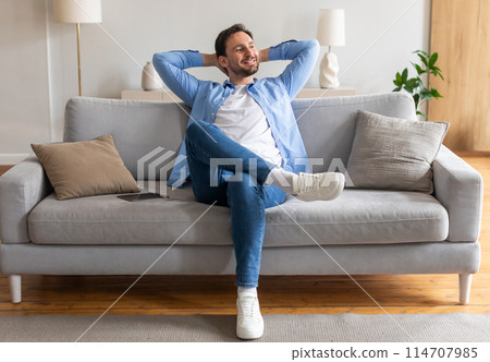 A man is seated on a couch, leaning forward with his hands cradling his head. He appears deep in thought or experiencing distress, showing signs of stress or contemplation. 114707985