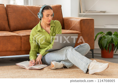 A focused young woman sits comfortably on the floor with her back against a couch, wearing headphones and using a laptop, possibly engaged in remote work or study A focused young woman sits comfortably on the floor with her back against a couch, wearing headphones and using a laptop, possibly engaged in remote work or study 114708005